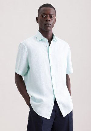 Mann trägt hellblaues kurzärmeliges Hemd mit Knöpfen und dunkle Hose, steht mit den Händen in den Taschen vor schlichtem Hintergrund.