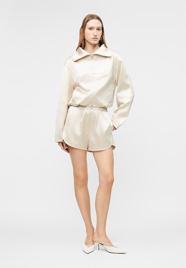 Femme portant un haut à manches longues en satin crème avec fermeture éclair et un short assorti, posant avec une main dans la poche et des mules blanches à talons.