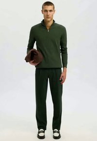 Sweater de fecho éclair verde escuro com colarinho de pé, combinado com calças da mesma cor. O modelo segura um item castanho e usa sapatos pretos e brancos.