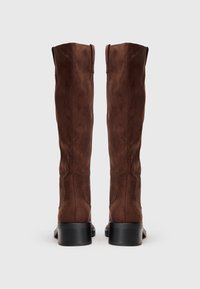 Botas de camurça castanhas acima do joelho com um design ajustado, bico quadrado e salto bloco baixo. Textura suave e detalhes de costura discretos.