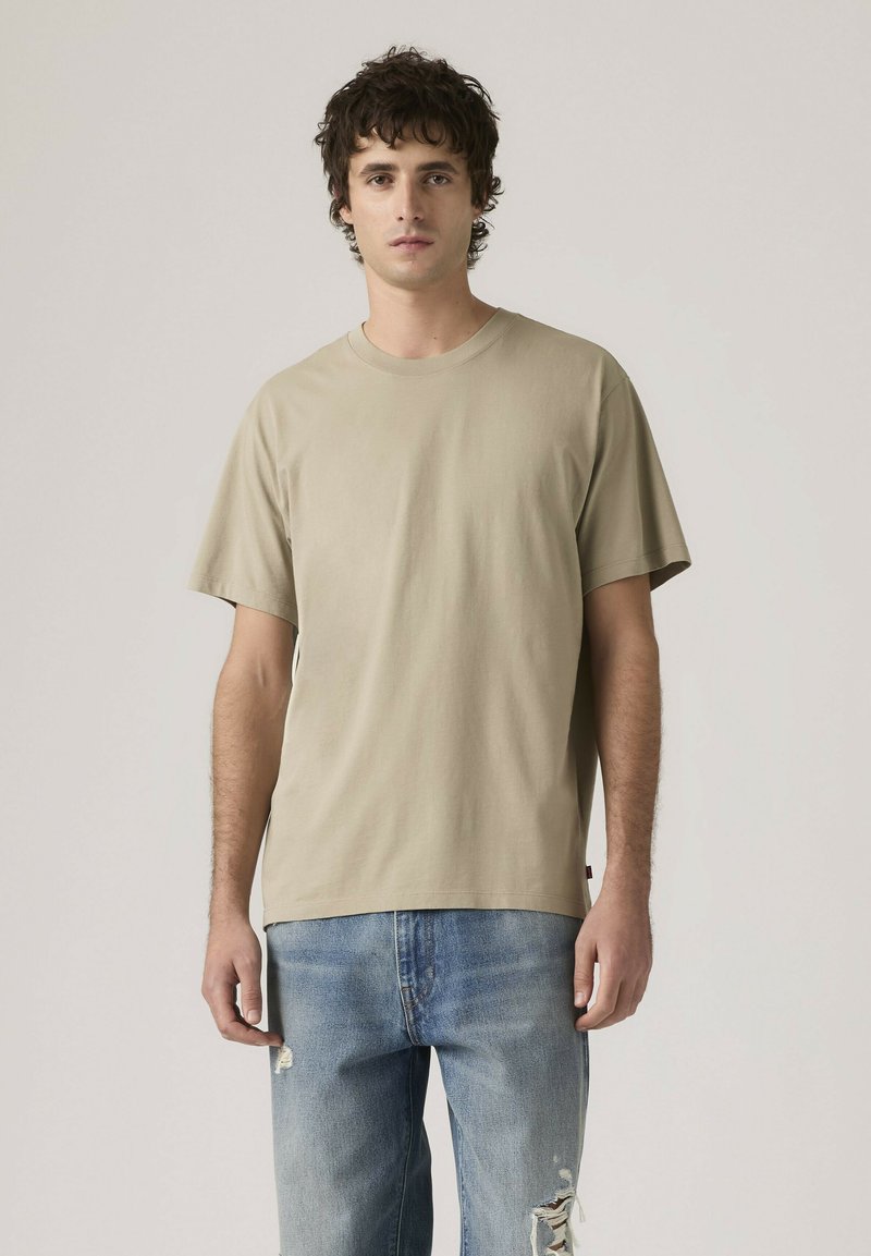 Giovane uomo con capelli ricci scuri che indossa una t-shirt beige a maniche corte e jeans strappati di colore azzurro chiaro, in piedi davanti a uno sfondo semplice.