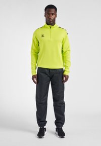 Ein Mann steht nach vorne blickend, trägt ein neongelbes Langarmshirt mit Viertel-Reißverschluss, schwarze Trainingshose mit elastischen Bündchen und schwarze Sportschuhe.