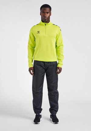 Mężczyzna stoi przodem, ubrany w neonowo żółtą bluzę z długim rękawem i zamkiem do ćwiartki, czarne spodnie dresowe z elastycznymi mankietami i czarne buty sportowe.