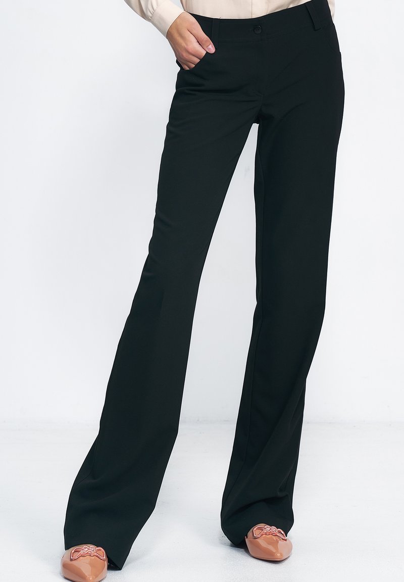 Osoba ubrana w czarne spodnie o prostym kroju i beżowe szpilki zakończone w szpic z detalem w postaci kokardy, z ręką w kieszeni, stoi na białym tle.