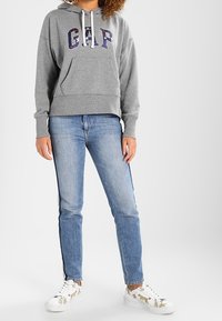 Felpa grigia con cappuccio e logo "GAP", tasca a marsupio, abbinata a jeans azzurri chiari e sneakers con stampa animale.