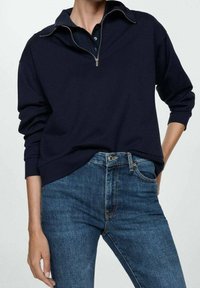 Sudadera de navy con cuello alto y media cremallera, con mangas anchas y un corte relajado, combinada con jeans ajustados azules.