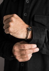 Juodas rankinis laikrodis su apvaliu ciferblatu ir sidabriniais akcentais, dėvimas ant struktūruoto juodo ilgomis rankovėmis marškinio. Įrengtas minimalizmo dizainu ir odiniu dirželiu.