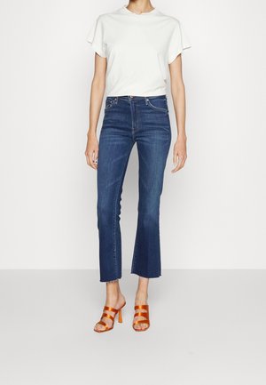 Personne portant un haut blanc à manches courtes, un jean bleu taille haute avec des ourlets évasés raccourcis, et des sandales à talons orange, se tenant devant un fond blanc uni.