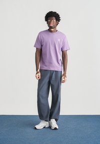 Μωβ βαμβακερό μπλουζάκι με κοντά μανίκια, γκρι φαρδιά παντελόνια και λευκά αθλητικά παπούτσια, σε απλό φόντο με μπλε δάπεδο.