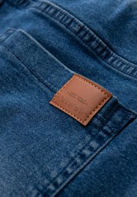 Pantalones vaqueros de mezclilla en un material azul oscuro, con un parche de cuero marrón cosido que dice "ambre est. 1985" cerca del bolsillo trasero.