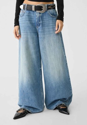 Wide-leg denim jeans in light blue with faded details. Features a black belt with a large silver buckle and studded accents. Pointed black shoes.