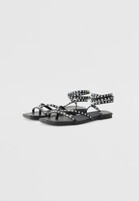 Des sandales plates noires comportant plusieurs lanières ornées de clous en argent, un design avec entre-doigts, et une semelle lisse et flexible.