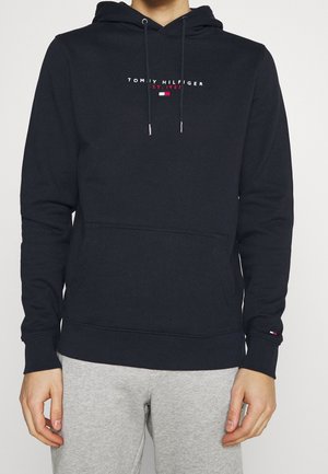 Person trägt einen schwarzen Tommy Hilfiger Hoodie mit Logotext und grauen Jogginghosen, steht mit entspannt an den Seiten hängenden Händen.