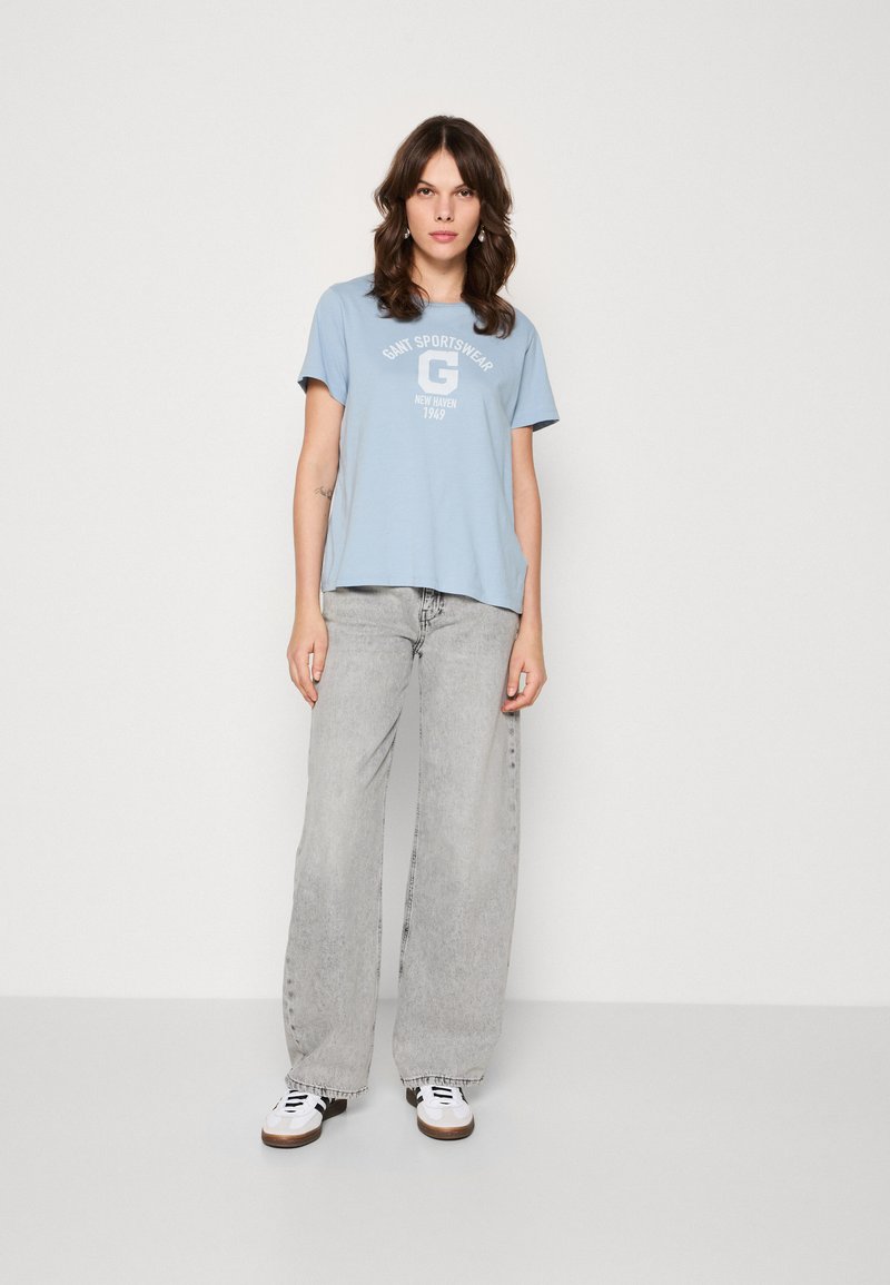 Kvinna står klädd i en ljusblå "Gant Sportswear" T-shirt, lösa grå jeans och vita sneakers mot en helt vit bakgrund.