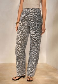 Mujer que lleva unos vaqueros de cintura alta y pierna ancha con estampado de leopardo en gris y beige, combinados con sandalias negras de tacón y una blusa sin mangas beige.