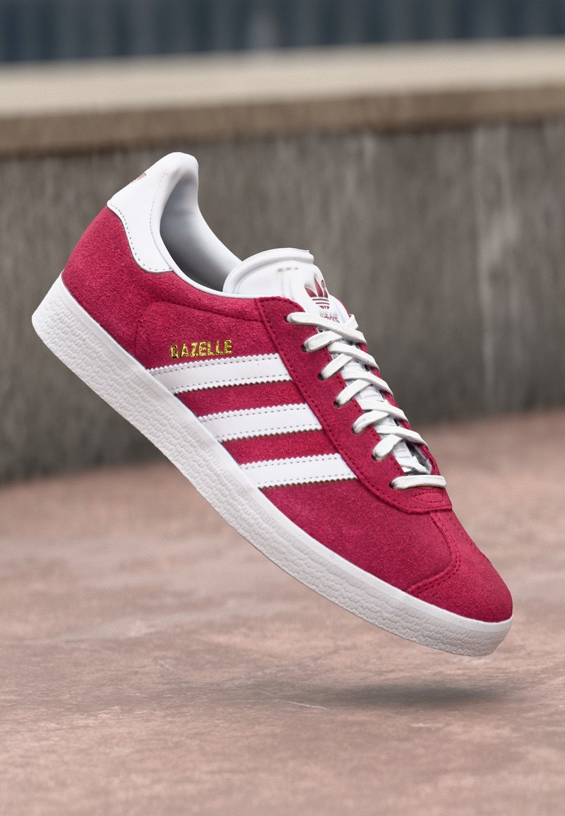 Roter Adidas Gazelle Sneaker mit weißen Streifen und Schnürsenkeln, weißem Fersenlaschen und strukturierter weißer Sohle, schwebend in der Luft vor Betonhintergrund.