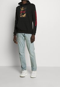 Sudadera negra con un gráfico frontal, texto rojo en las mangas, combinada con jeans de un tono azul claro y zapatillas blancas. Tejido robusto, corte relajado.