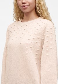 Jeune femme aux longs cheveux blonds bouclés porte un pull rose clair avec une texture tricotée en relief à petites boules sur les épaules et les manches.