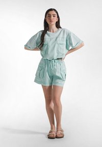 Miętowozielony, oversize'owy top z dwoma przednimi kieszeniami oraz pasujące szorty ze sznurkiem w talii i bocznymi kieszeniami. Modelka ma na sobie jasne sandały.