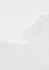 Camiseta blanca de algodón con cuello redondo. Presenta una textura suave y un dobladillo cosido. La etiqueta dice "Pier One" en el interior cerca del cuello.