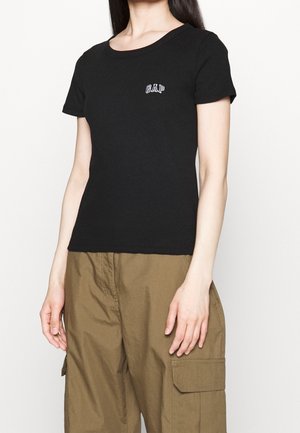 Vrouw die een zwart korte mouwen GAP T-shirt en een bruine cargobroek draagt tegen een effen lichte achtergrond.