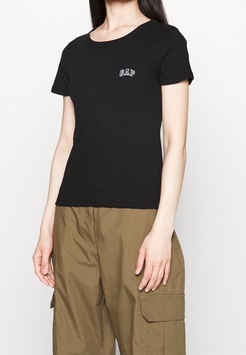 Femme portant un t-shirt noir à manches courtes GAP et un pantalon cargo marron sur un fond clair uni.
