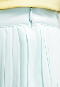 Gonna plissettata azzurra chiara con una texture morbida, dotata di una zip discreta e una vita, mostrata parzialmente contro una maglia gialla.