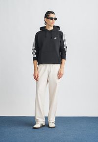 Μαύρο φούτερ με λευκό σχέδιο τριών ρίγων και λογότυπο, σε συνδυασμό με ανοιχτό μπεζ παντελόνι. Το μοντέλο φορά γυαλιά ηλίου σε σκούρο χρώμα και λευκά αθλητικά παπούτσια.