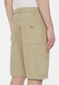 Beige työshortsit kestävästä kankaasta, useilla taskuilla, takana logopatch ja vahvistetut saumat lisäämään kestävyyttä.