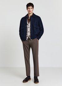 Giacca blu navy in misto lana con tasche frontali, indossata sopra un cardigan beige, abbinata a pantaloni marroni su misura e scarpe scure.
