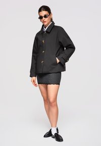 Femme portant une veste noire boutonnée, une mini-jupe grise à côtes avec bordure en dentelle, des mocassins noirs avec des chaussettes blanches et des lunettes de soleil noires fines.