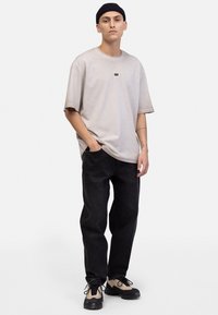 Beige oversized t-skjorte med en liten svart logo på brystet, kombinert med mørk denim jeans og svarte sneakers med beige detaljer.