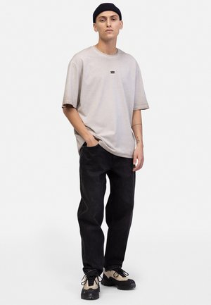 Bēša oversized t-krekls ar mazu melnu logotipu uz krūts, apvienots ar tumša denim šķiedru džinsiem un melniem sporta apaviem ar bēšiem akcentiem.