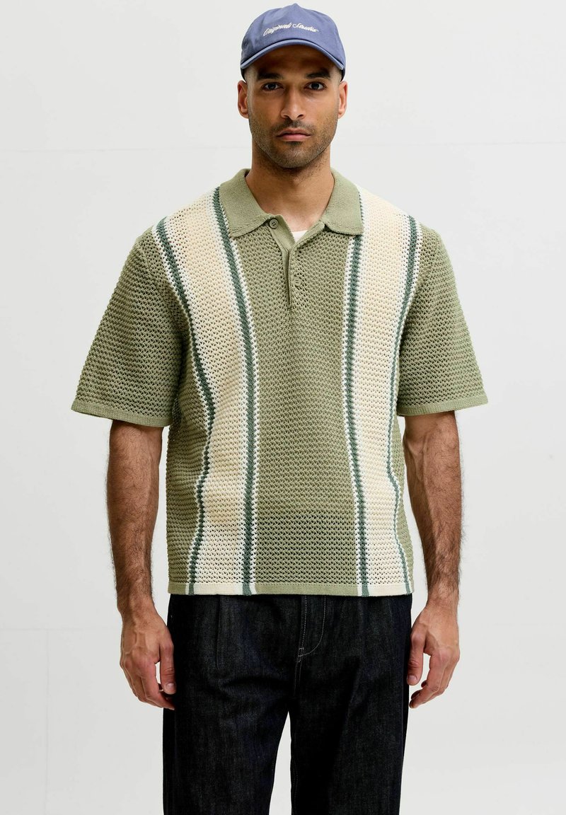 Homme portant un polo en maille à manches courtes rayé vert et crème et une casquette bleue, debout devant un fond clair uni.