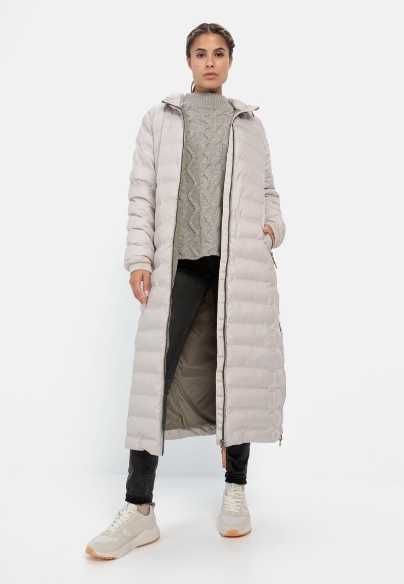 Gevoerde lange jas in lichtbeige met een capuchon, geribbelde textuur, ritssluiting aan de voorkant en zakken, gecombineerd met een grijs gebreid vest en zwarte jeans.