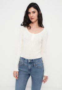 Κρεμ πουκάμισο με μακριά μανίκια, ριμπ υφή και στρογγυλή λαιμόκοψη. Συνδυάζεται με ανοιχτό μπλε στενά τζιν με κλασικό σχεδιασμό πέντε τσεπών.