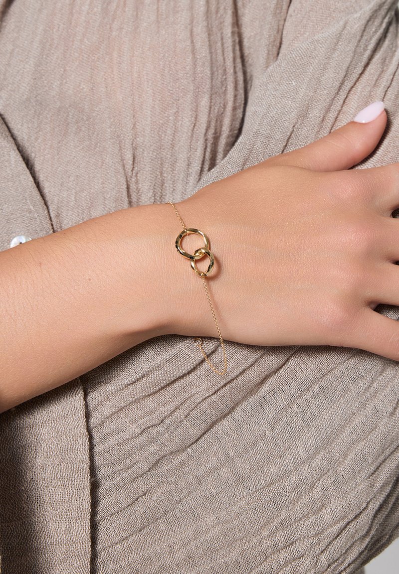 Złota bransoletka z przeplatającymi się okrągłymi ogniwami, delikatny łańcuszek, noszona na dłoni na tle teksturowanej brązowej tkaniny.
