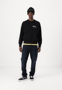 Sort sweatshirt med hvid grafisk design, gul undertrøje, mørkeblå jeans, sort-hvide sneakers og en sort hue.