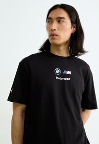 Sort bomulds T-shirt med BMW og M Motorsport-logoer på brystet, korte ærmer og subtile lyserøde accenter langs siderne.