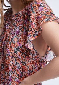 Bluse aus transparenter, floral gemusterter Stoff in Orange-, Violett- und Grüntönen. Verfügt über kurze, rüschierte Ärmel und einen runden Halsausschnitt mit ausgeschnittenem Detail.