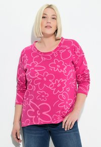 Różowa bluzka na długi rękaw z kwiatowym wzorem w ciemniejszym odcieniu różu. Materiał wydaje się miękki, z zaokrąglonym dekoltem i luźnym krojem.