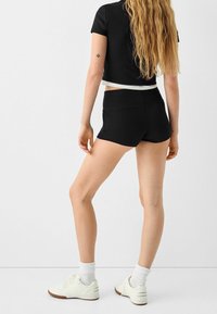 Femme aux longs cheveux blonds portant un crop top noir à manches courtes, un short noir, des chaussettes blanches et des baskets blanches, se tenant de dos.