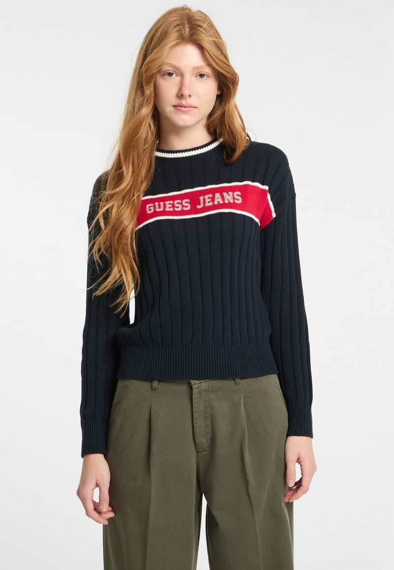 Marinblå stickad tröja med ribbad textur, med ett tydligt röd och vit logoband som läser "GUESS JEANS" över bröstet.