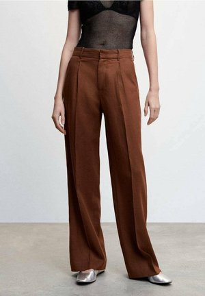 Kvinde iført brune højtaljede, vidde bukser, sort gennemsigtig top og sølvfarvede flade spidse sko, stående mod en ensfarvet baggrund.