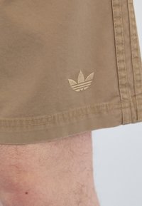 Rudi šortai iš minkštos medžiagos, su mažai išsiuvinėtu trimis juostelėmis logotipu ant krašto, matomais siuvimo detalėmis palei kraštą.