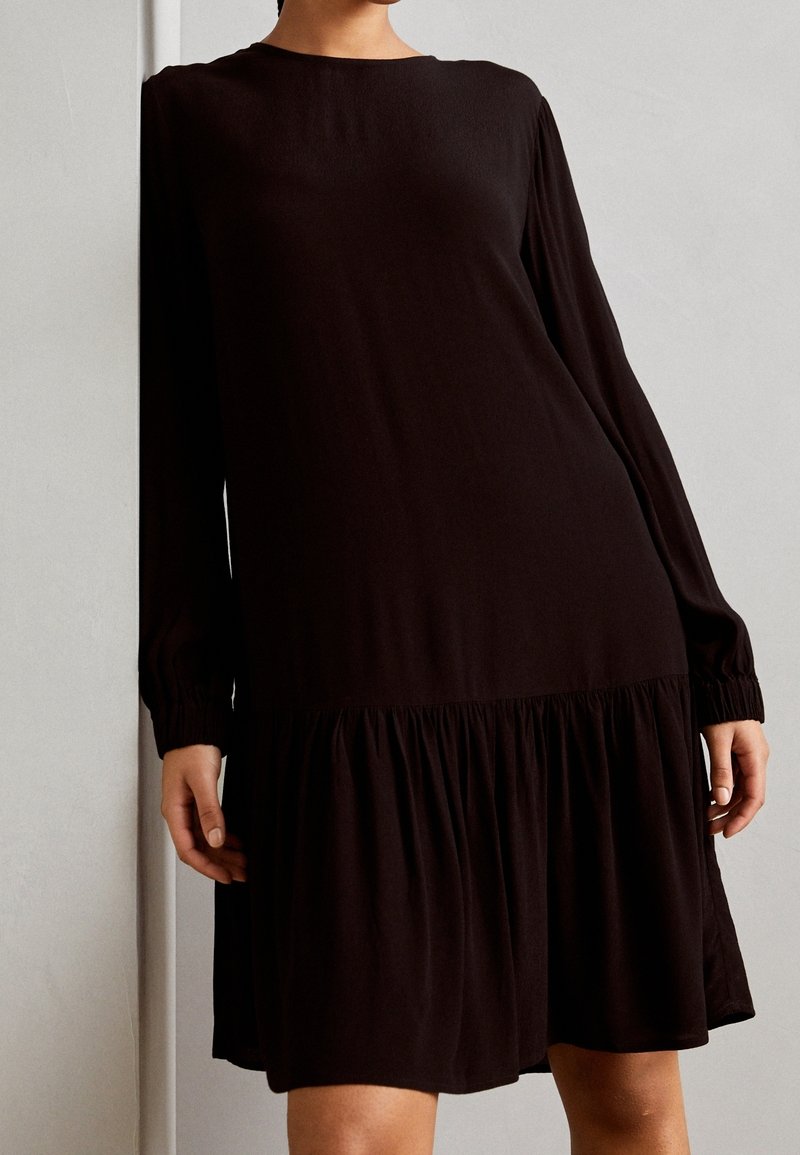 Vestido negro de manga larga con parte inferior acampanada, hecho de tela suave. Presenta puños fruncidos y un clásico escote redondo.