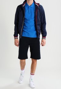 Marinefarbene Zip-Up-Jacke mit rot-weißen Streifenakzenten, leuchtend blauen Poloshirt, schwarzen Shorts, weißen Sneakern und rot-gestreiften Socken.