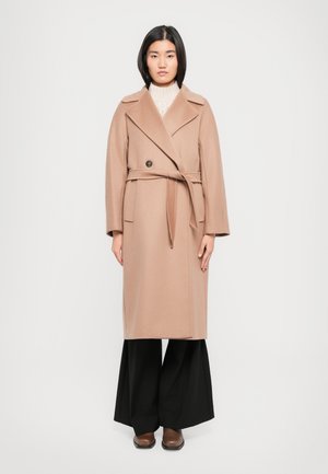 Cappotto in lana marrone chiaro con vita cinturata, revers a punte e chiusura con un solo bottone, abbinato a pantaloni neri a gamba larga e scarpe marroni.