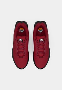 Red sneakers with a textured upper, black sole, and round logos on the tongue. Features a lace closure and curved midsole design.