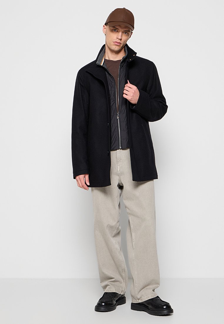 Manteau noir en mélange de laine avec un col texturé, superposé à une veste matelassée grise, assorti à un pantalon beige et des chaussures noires.